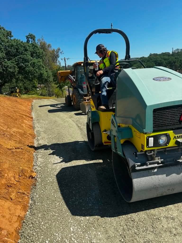 Two workers ride a yellow road roller paving a new asphalt road beside an orange dirt embankment.