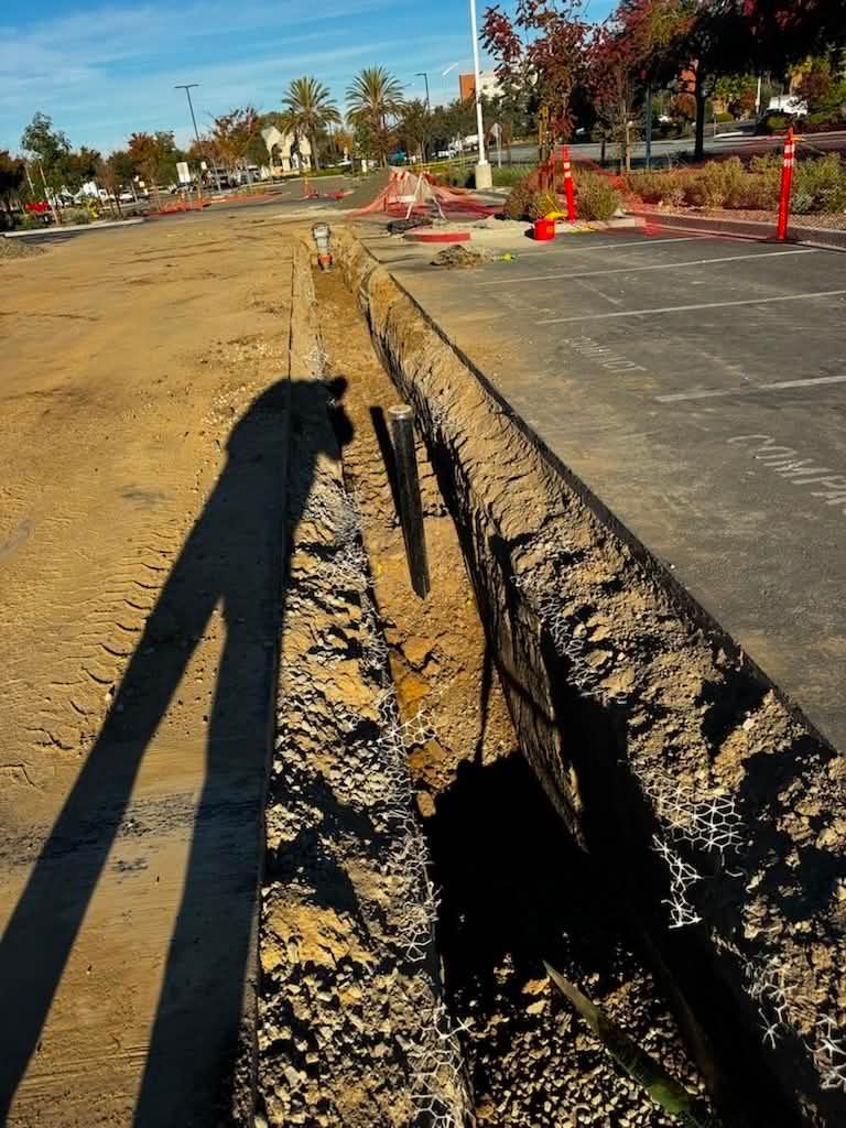 Road construction site with a deep trench beside a paved lane and orange safety cones in bright sunlight