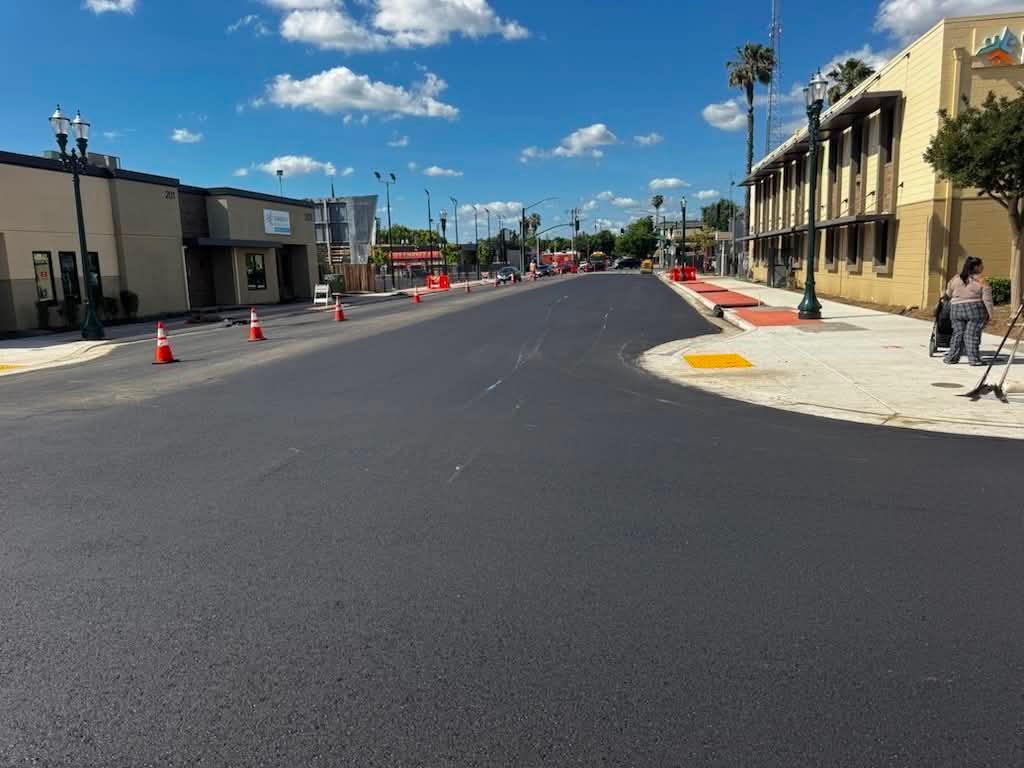 Freshly paved street with orange cones, buildings on both sides, and a blue sky with clouds.