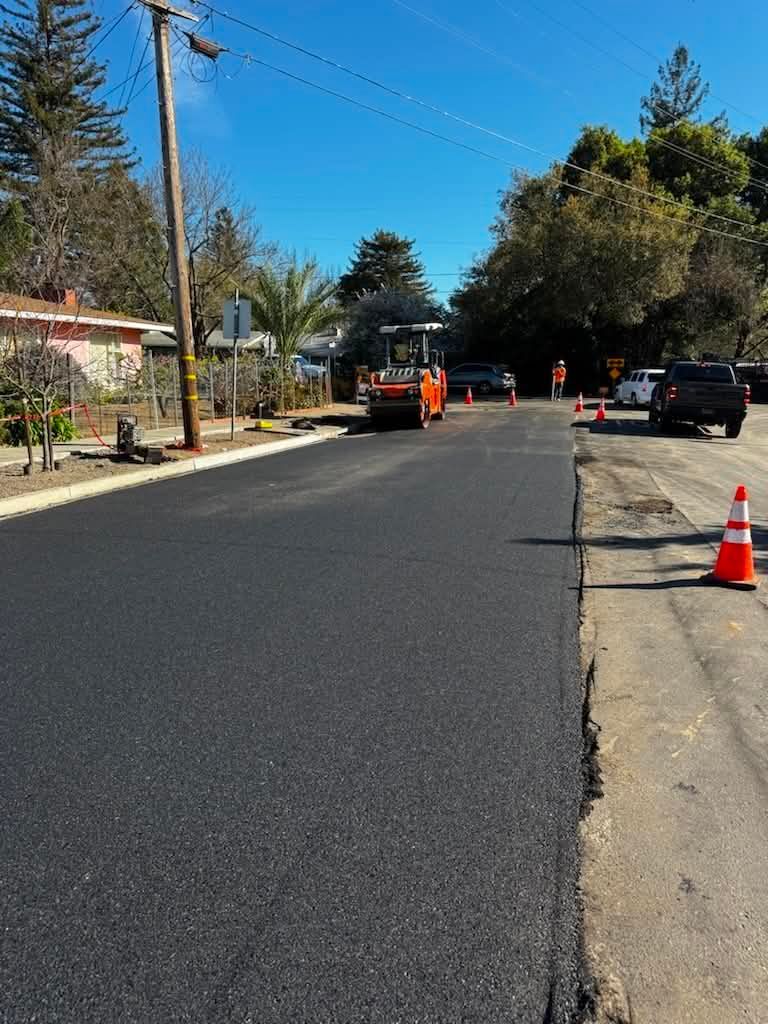 Roadwork on a quiet residential street with orange cones, a worker, and parked cars under a clear blue sky