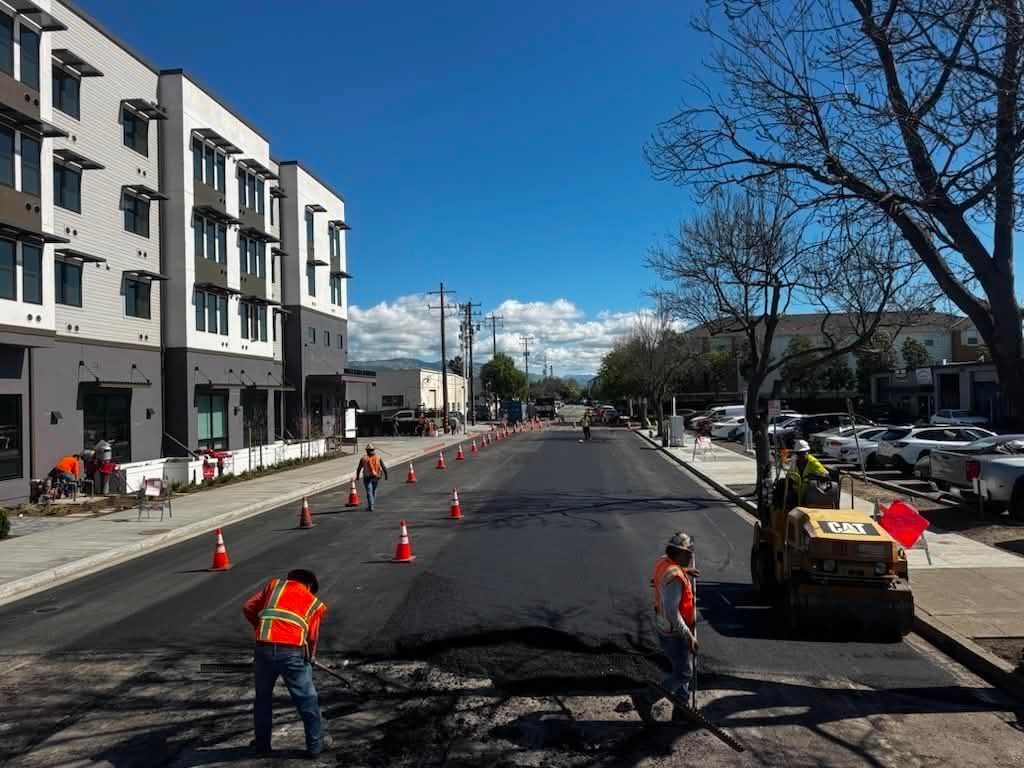 Road construction with cones, workers, and a steamroller on a city street under a blue sky