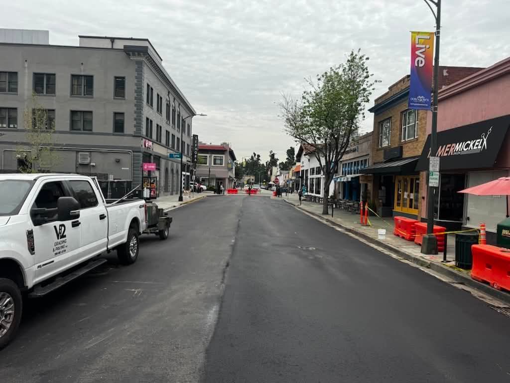 Quiet downtown street with parked cars, storefronts, and a white truck on the left