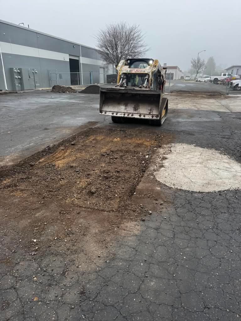 Skid steer in a muddy parking lot beside a building, with a scraped patch of pavement nearby.
