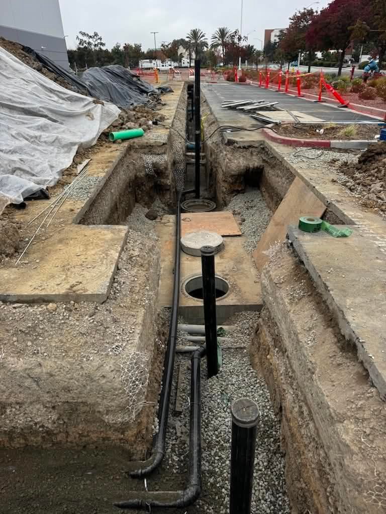 Construction trench with exposed utilities and pipes, bordered by gravel and barriers along a road.