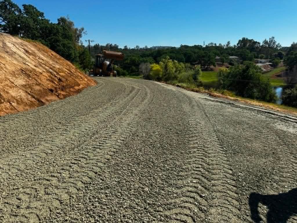Gravel road under construction beside a dirt embankment and green trees on a sunny day