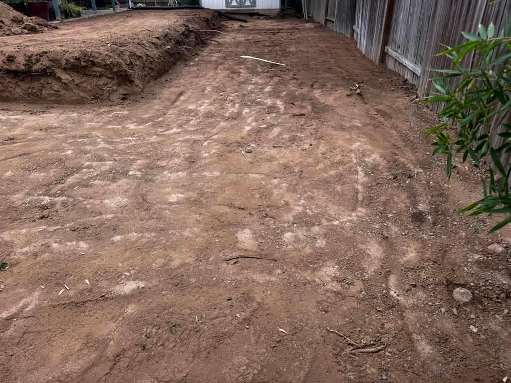Dirt path or driveway under construction, with uneven soil and a fence along the right side.
