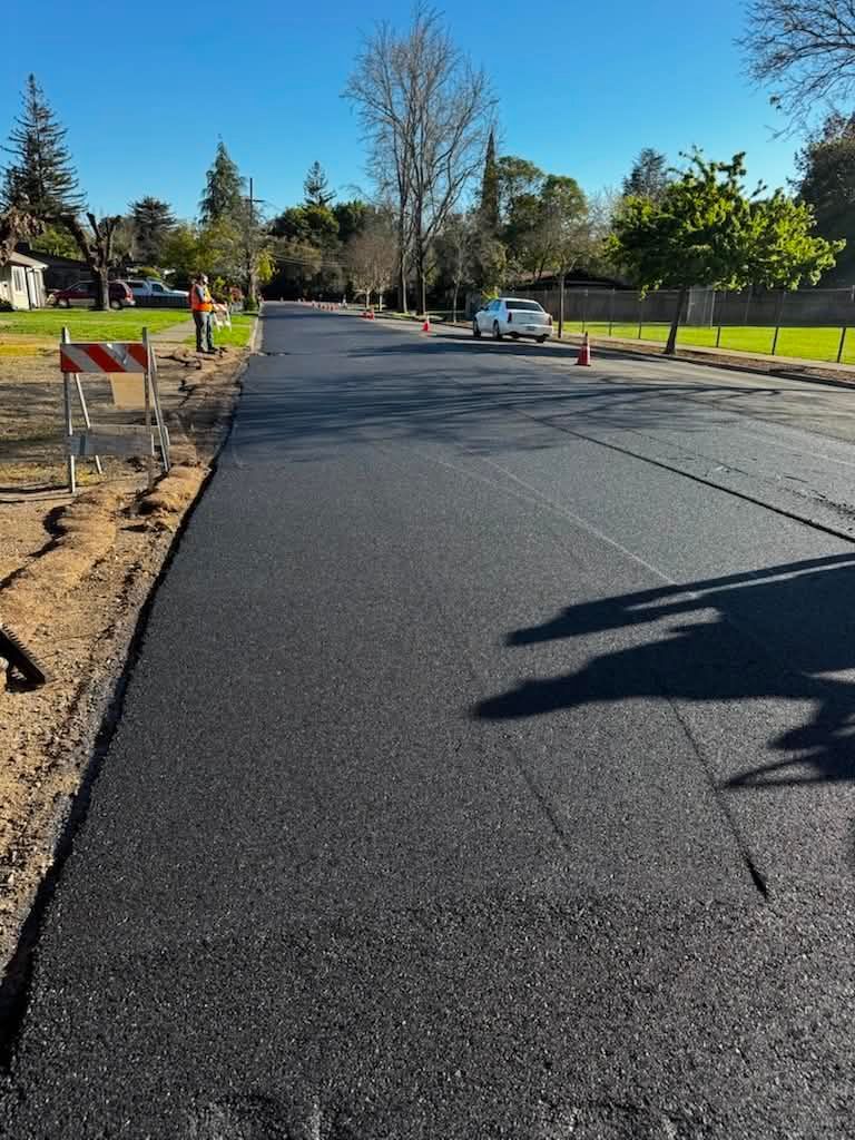 Residential street with fresh black pavement, orange cones, and a car parked ahead on a sunny day