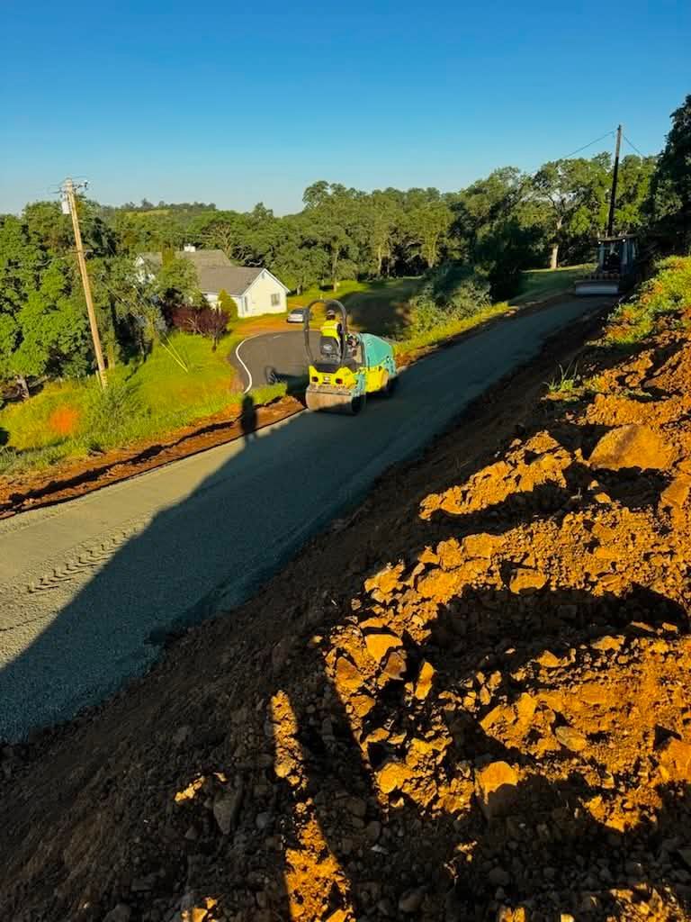 Road construction on a hillside with an excavator, dirt embankments, and houses in the distance.