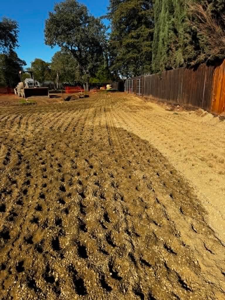 Plowed field with rows of soil beside a wooden fence and trees under a clear blue sky