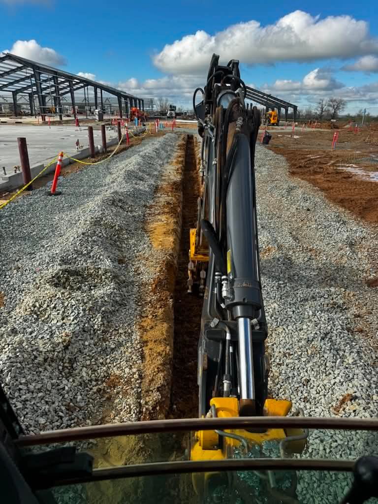 Excavator arm over gravel trench at a construction site with orange cones and metal framework under cloudy sky
