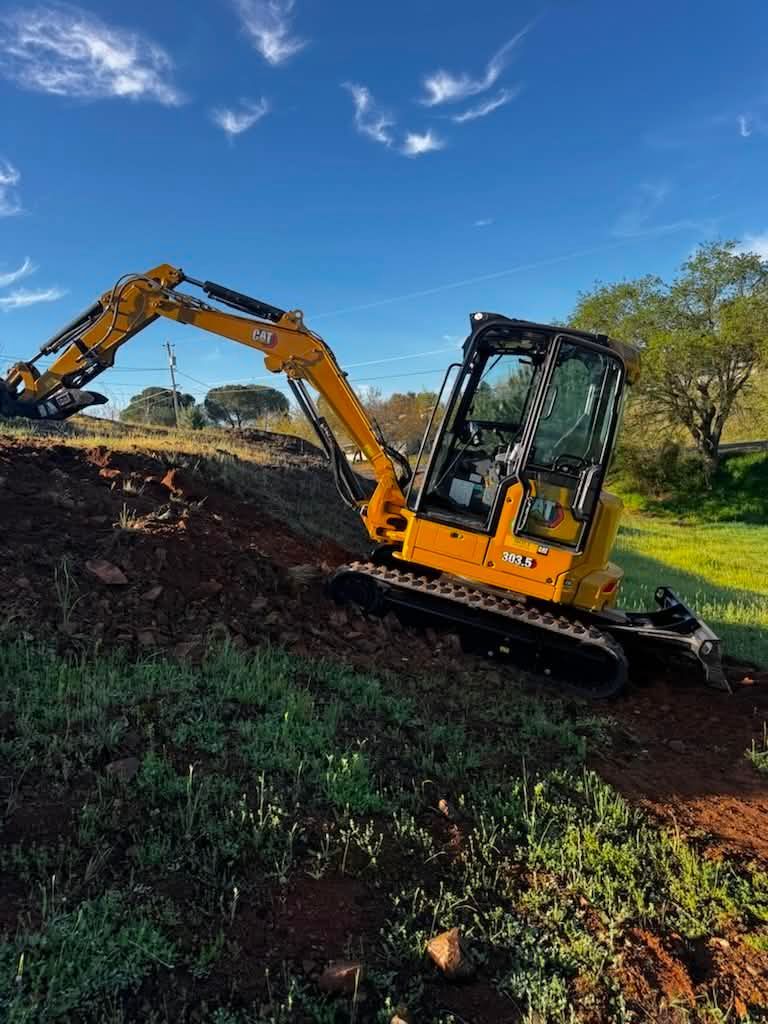 Yellow excavator digging on a dirt slope under a blue sky, with green grass and trees nearby