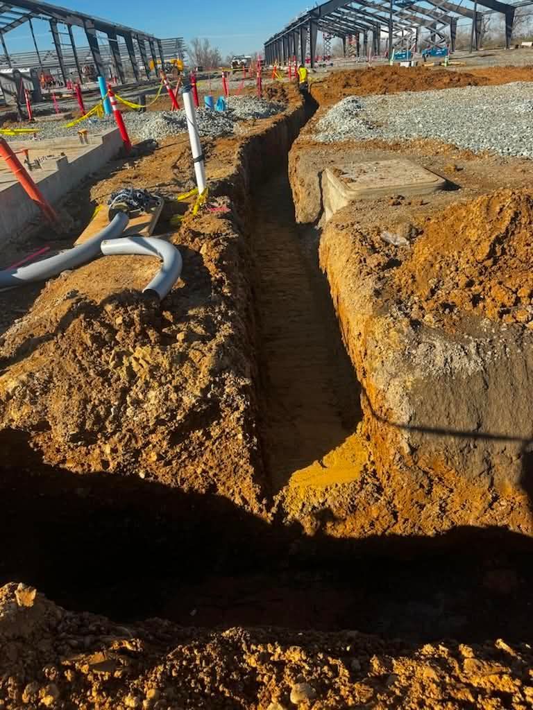 Trench dug beside a concrete foundation at a construction site, with orange safety markers and pipes.