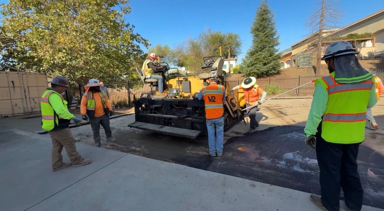 Road crew in safety vests paving asphalt on a residential street, with a paver machine and workers guiding it.