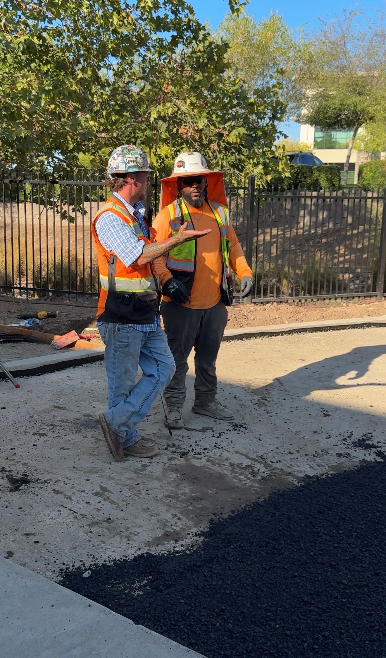 Two construction workers in safety vests and hard hats stand on a road paving site.