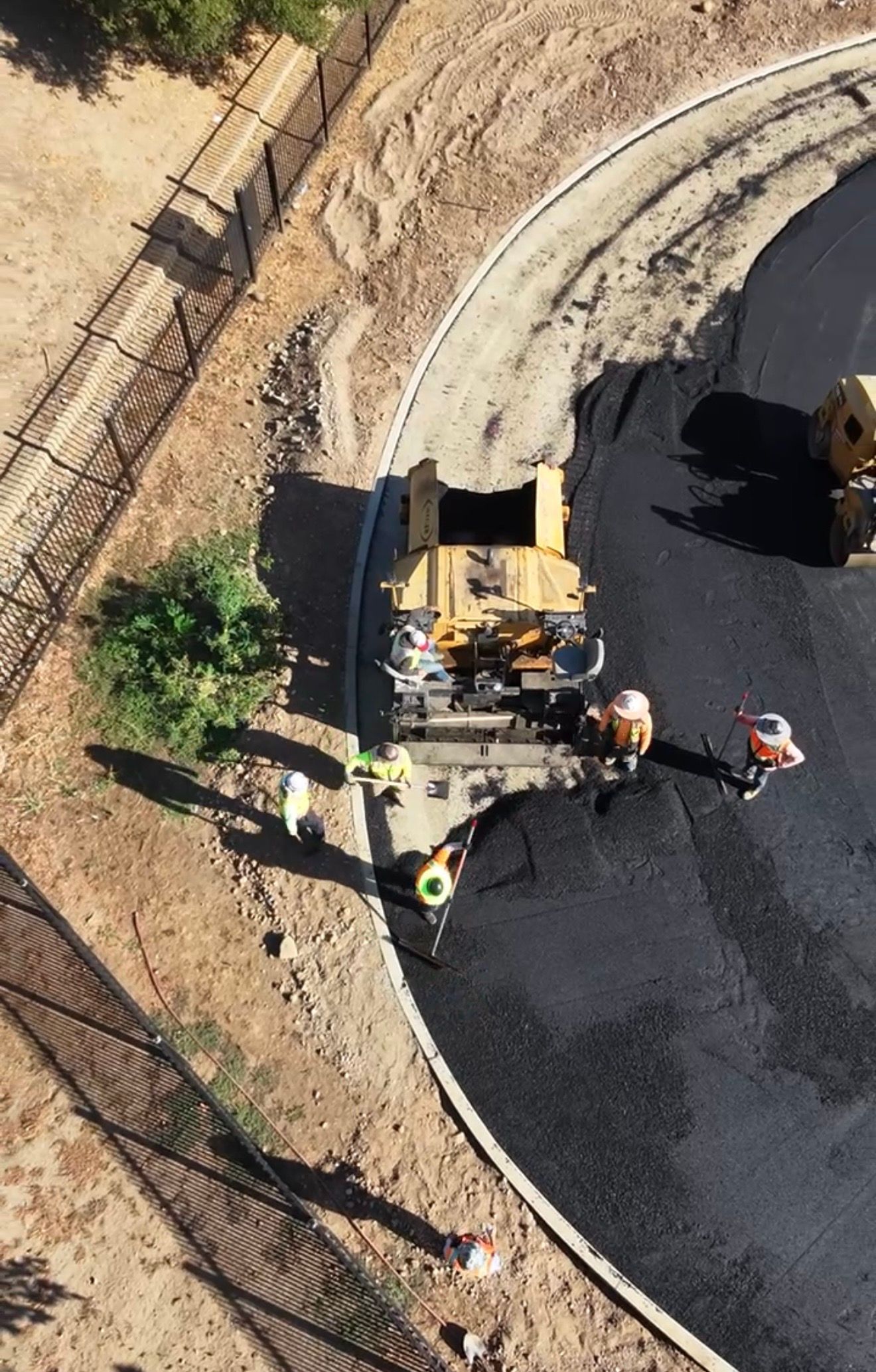 Road workers paving a curved road with a yellow asphalt machine beside a dirt shoulder.