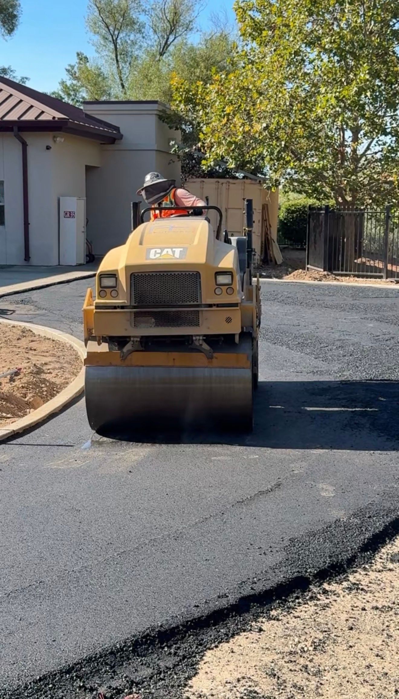 Yellow road roller paving a new black asphalt driveway beside a house and trees.