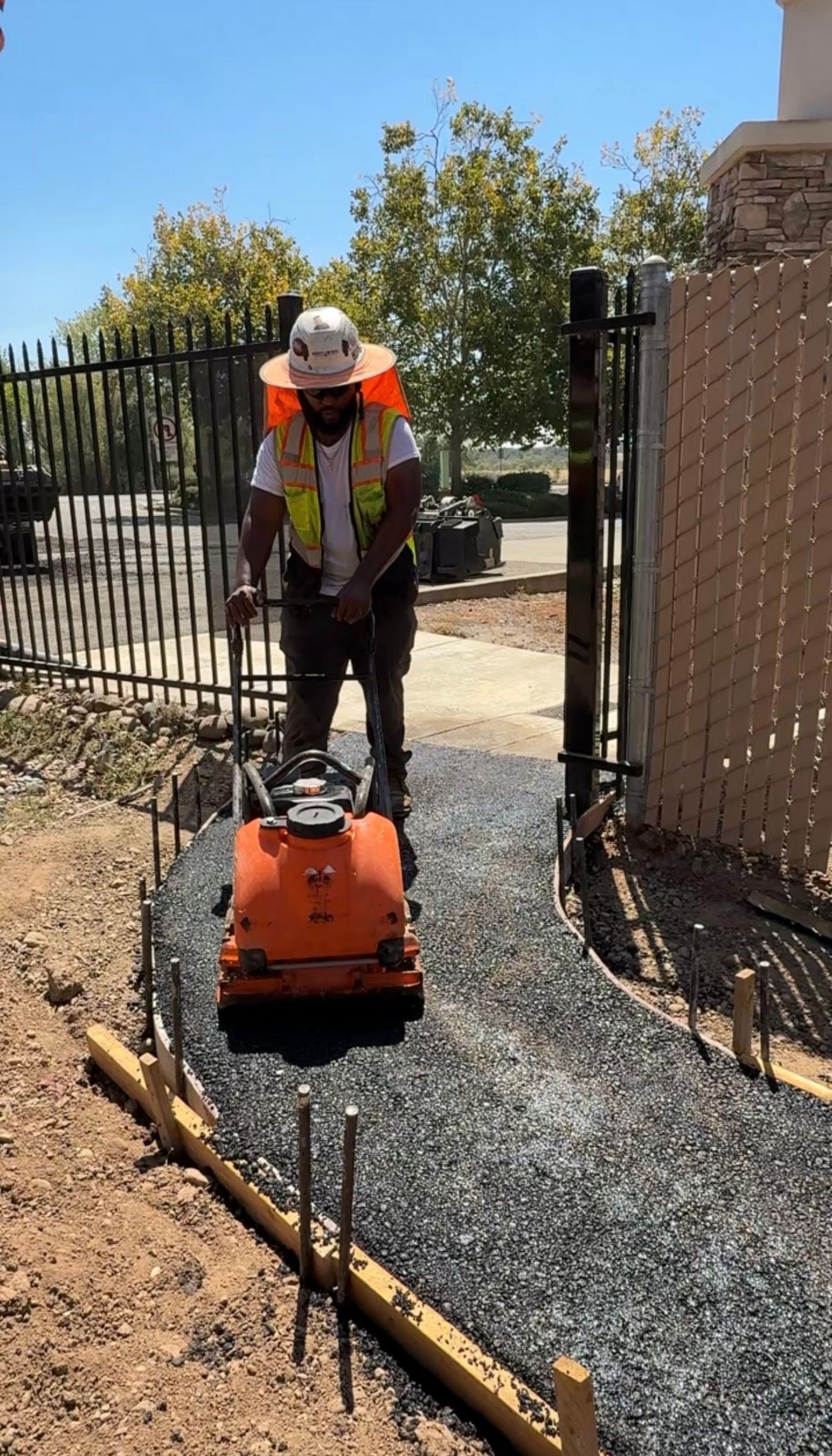 Construction worker operating orange plate compactor on gravel path beside a fence and dirt trench