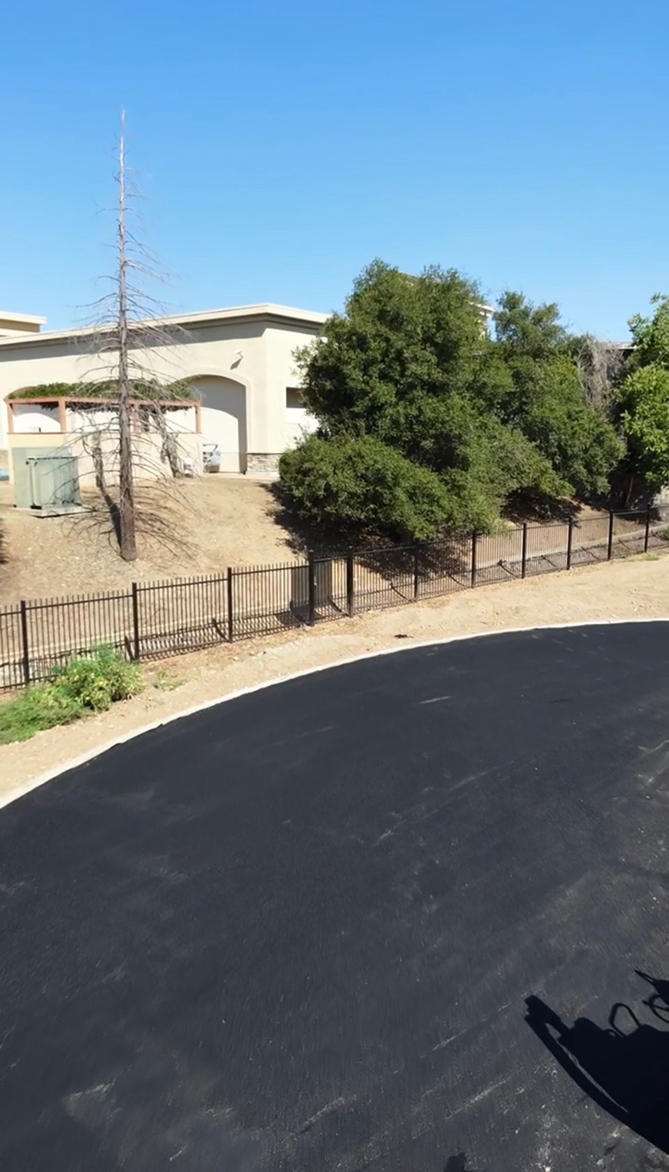 Curved black asphalt road beside a beige building and green trees under a clear blue sky