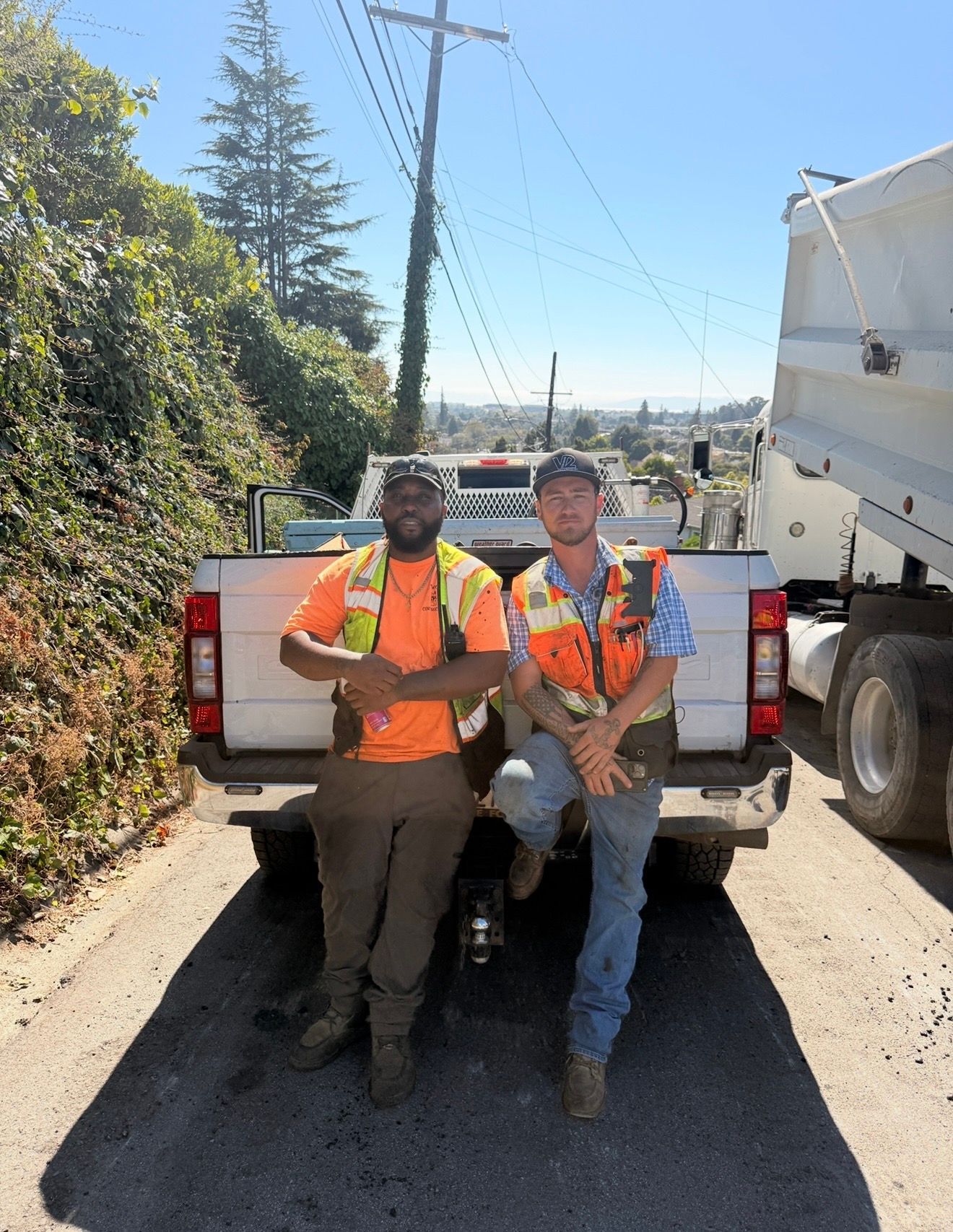 Two utility workers in orange vests sit on a pickup tailgate beside a roadside work truck.