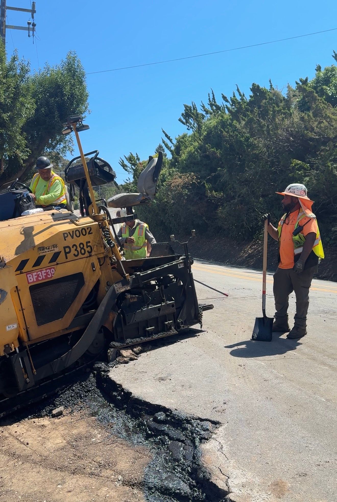 Road repair crew operating asphalt paver beside a fresh black road patch on a sunny street