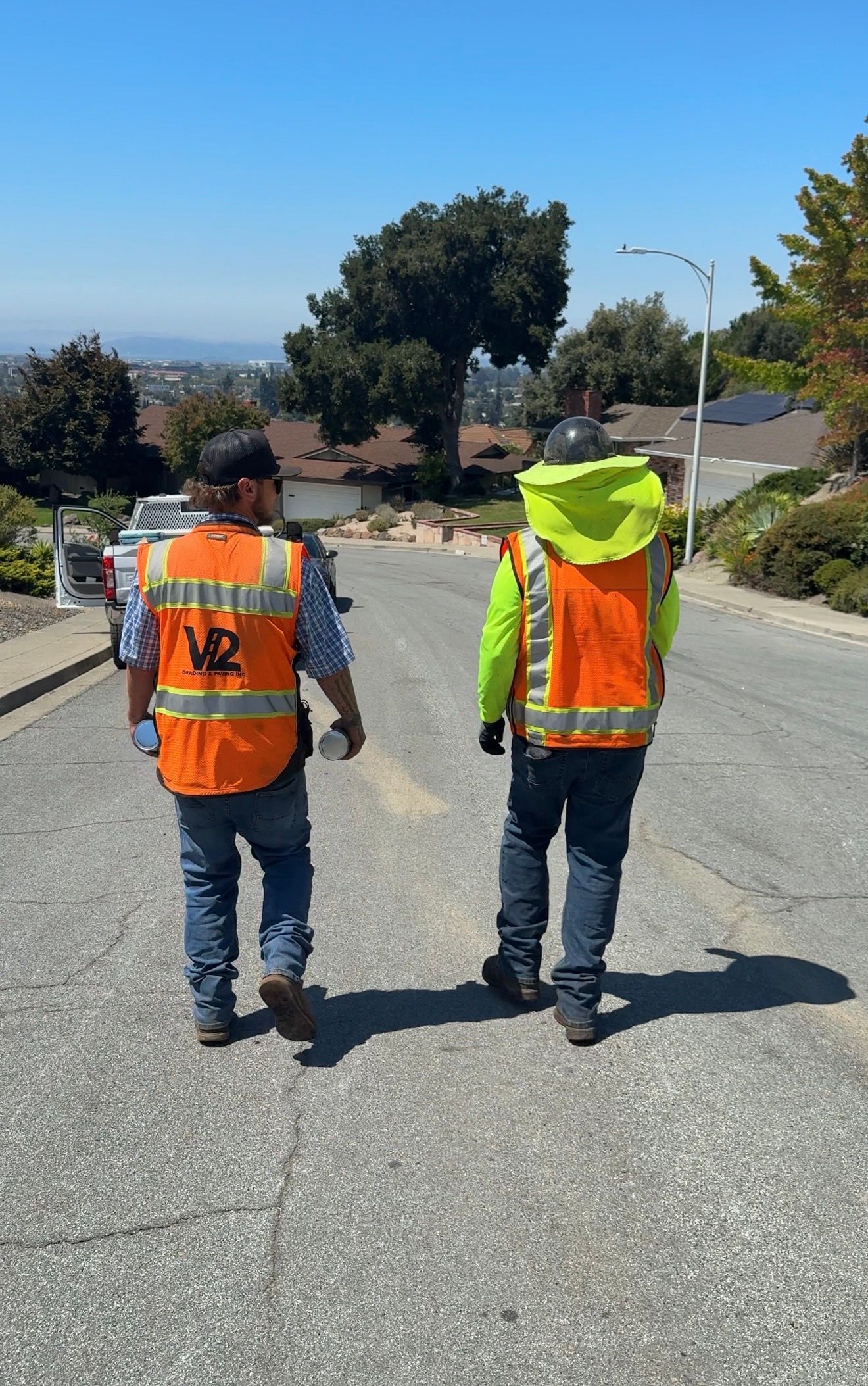 Two road workers in orange safety vests walking away on a sunlit gravel road, one carrying a backpack.