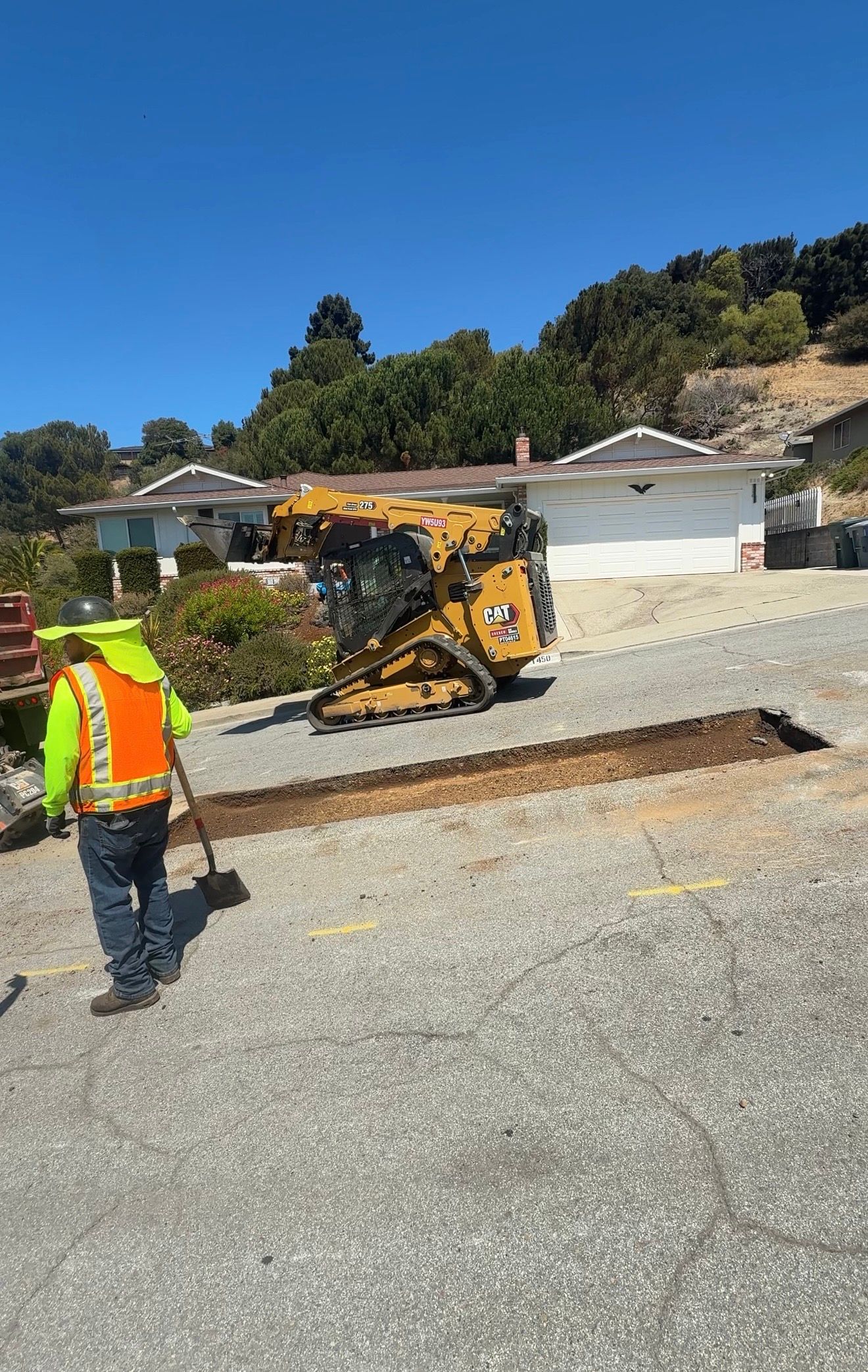 Road worker in neon vest beside yellow excavator on a gravel street under a clear blue sky