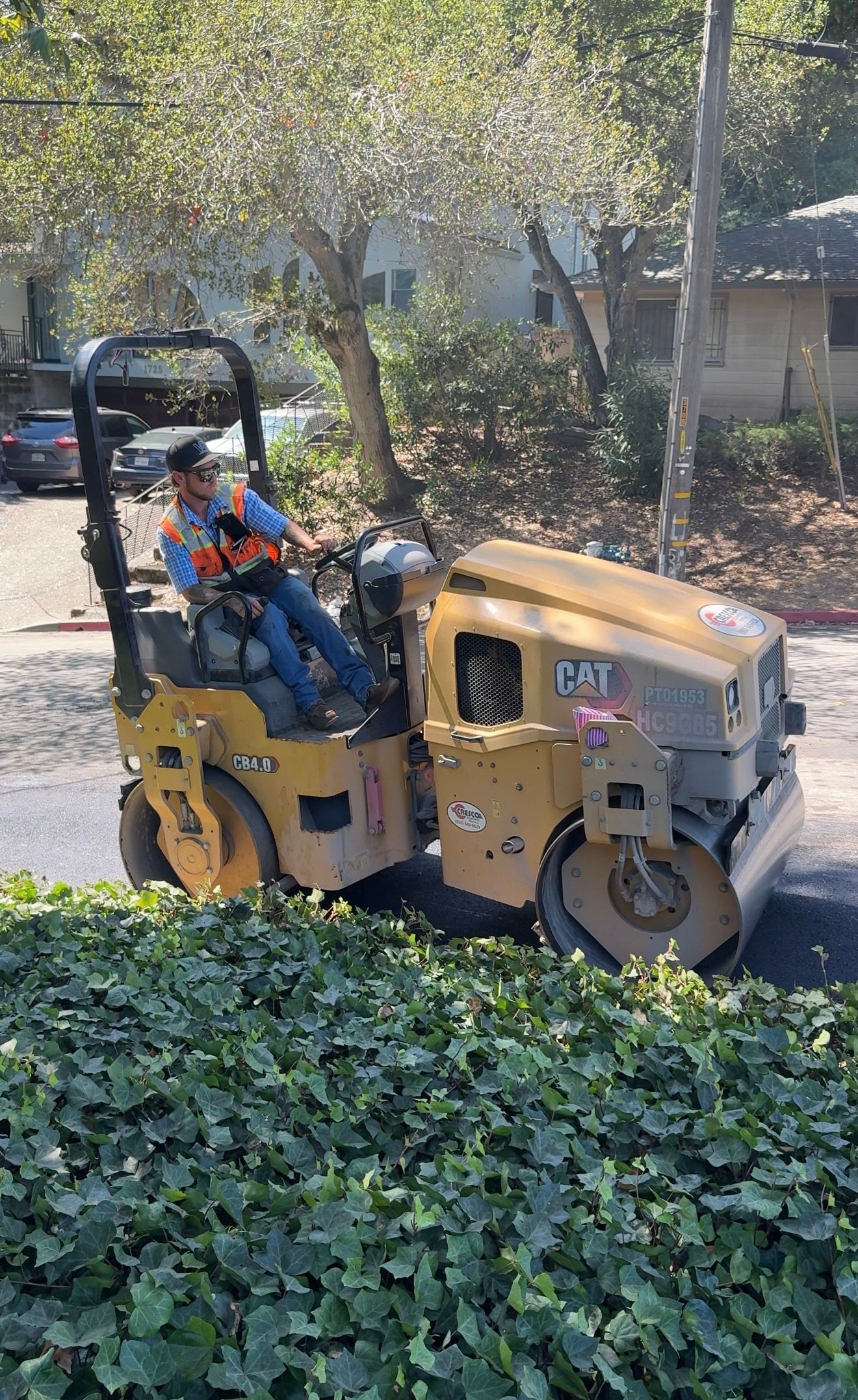 Yellow tractor operating in a leafy area, with a driver wearing an orange vest and hard hat.