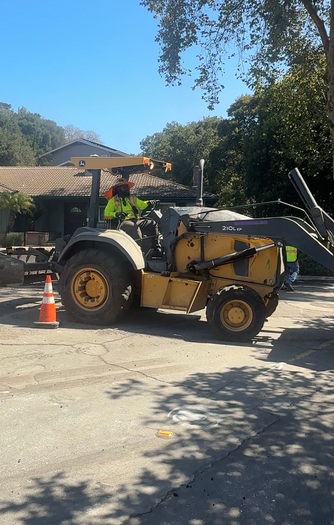 Yellow tractor parked on a sunny street beside an orange traffic cone and trees