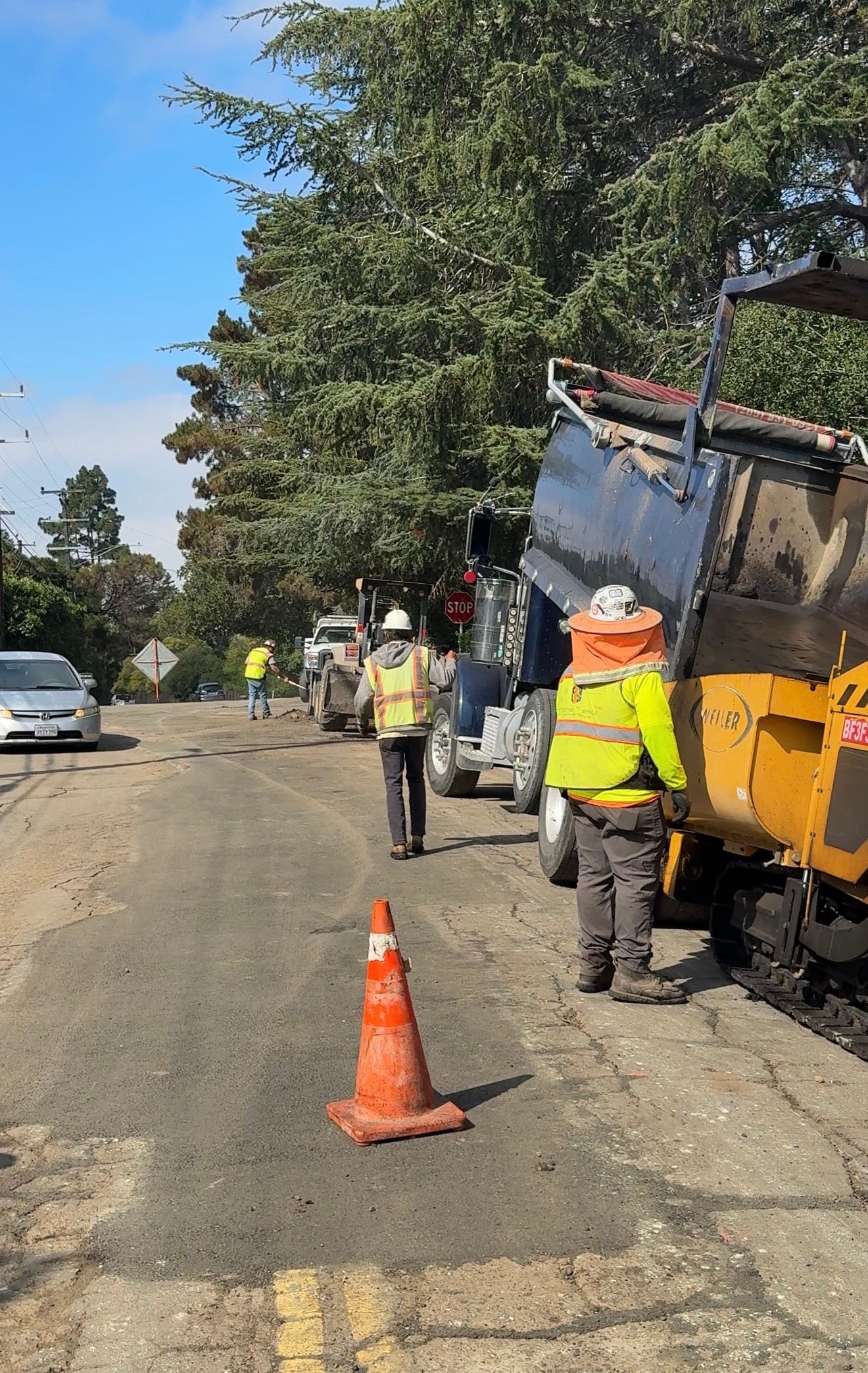 Roadwork on a gravel roadside with workers in yellow vests, traffic cones, and a yellow truck.