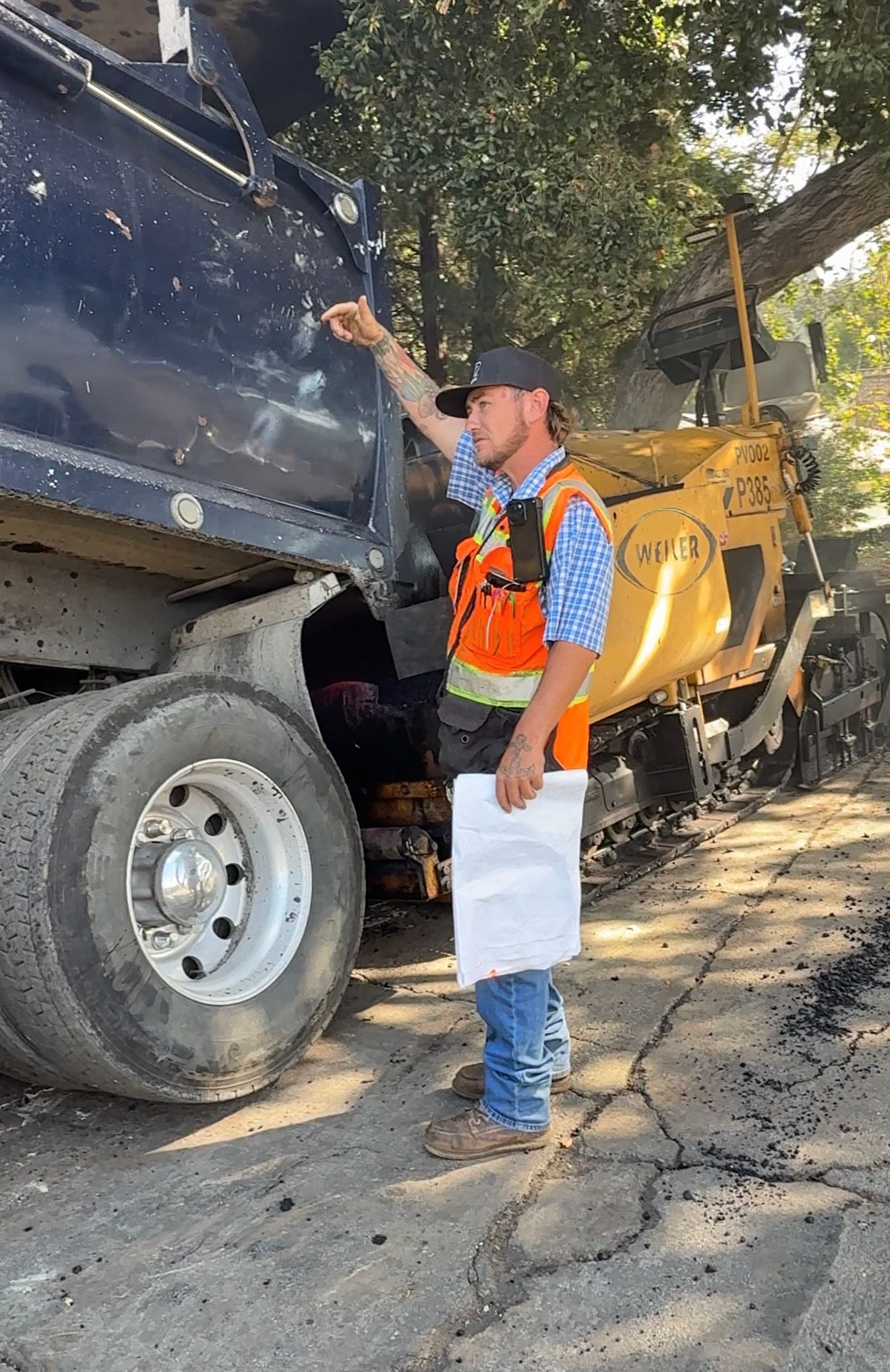 Worker in orange vest inspecting a black dump truck beside a yellow excavator on a dirt road.