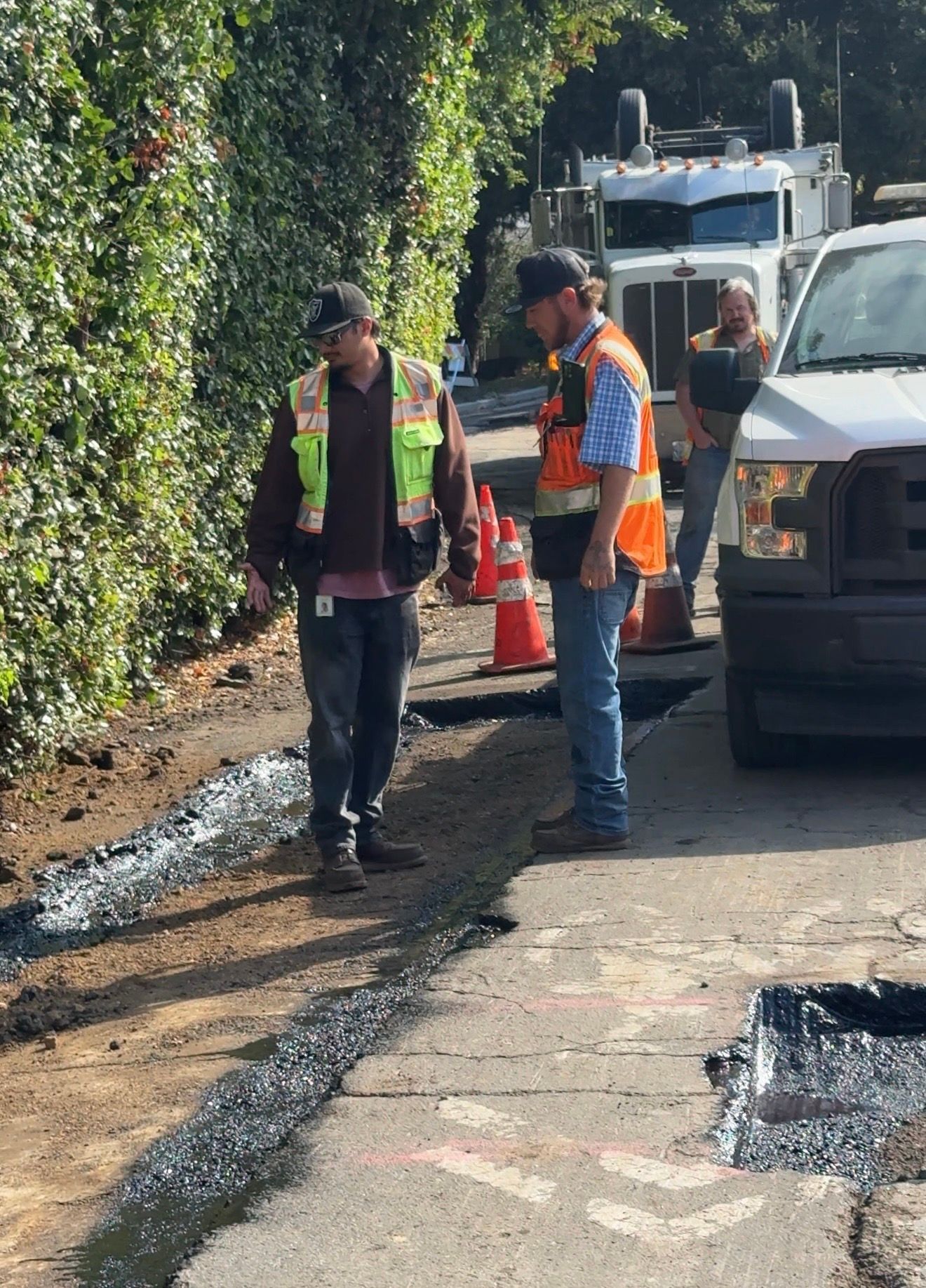 Two road workers in high-visibility vests inspect a roadside trench beside traffic cones and parked vehicles.