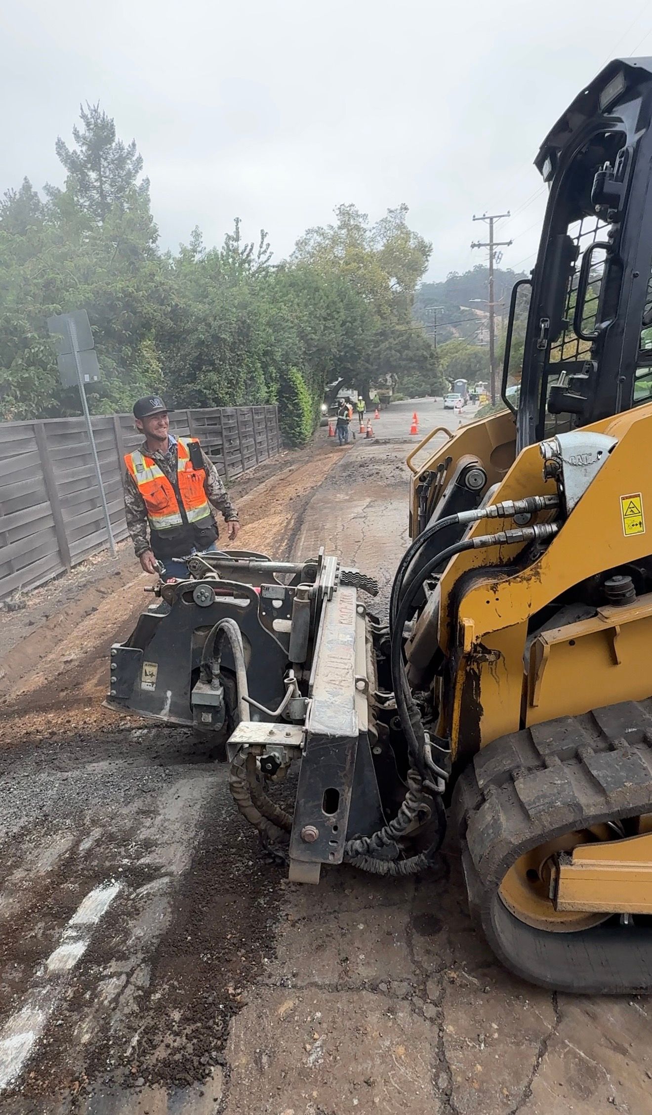 Yellow excavator clearing debris on a muddy road while a worker in orange stands nearby.