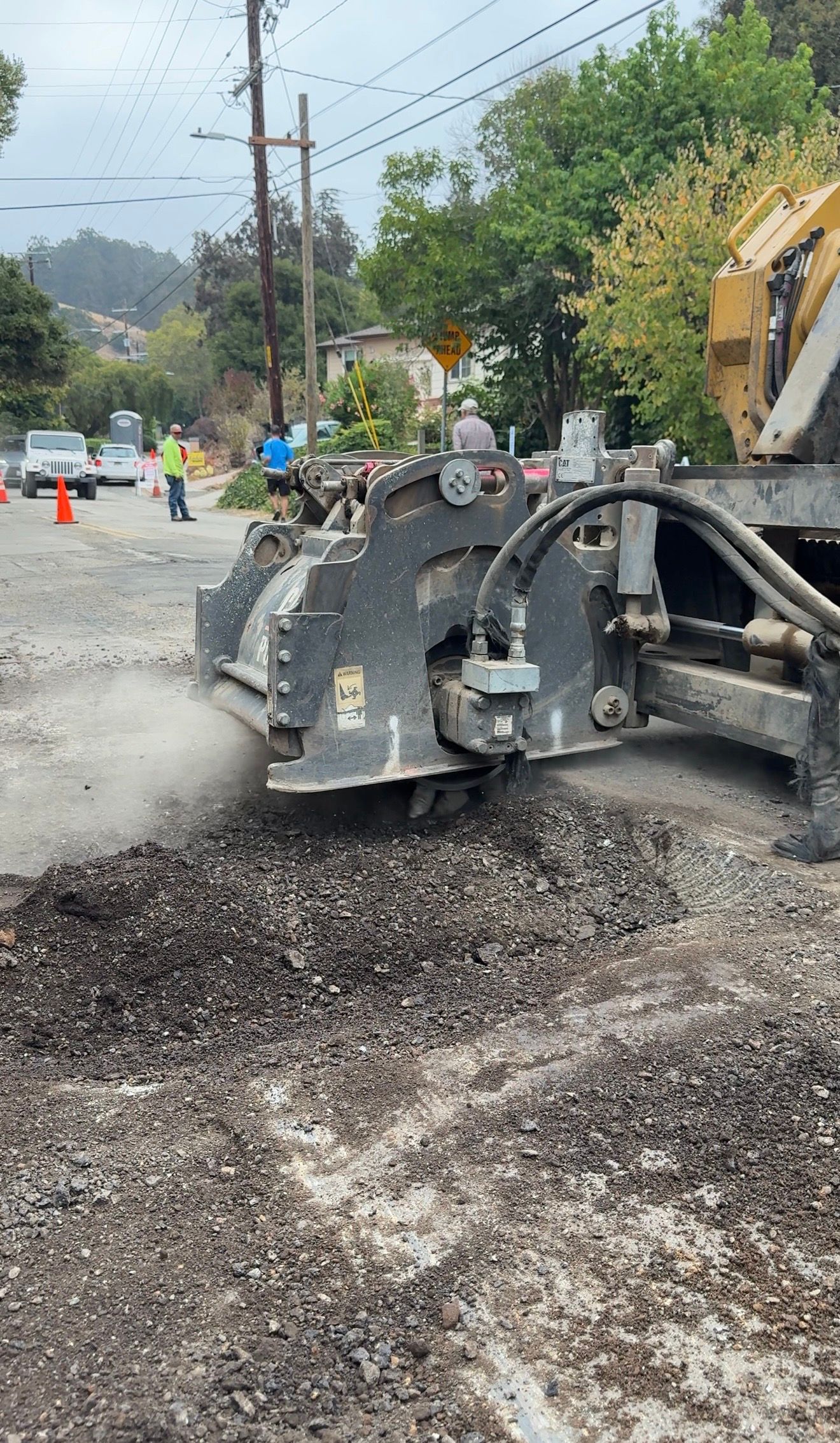 Road paving machine spreading dark asphalt on a wet street beside orange cones and roadside trees