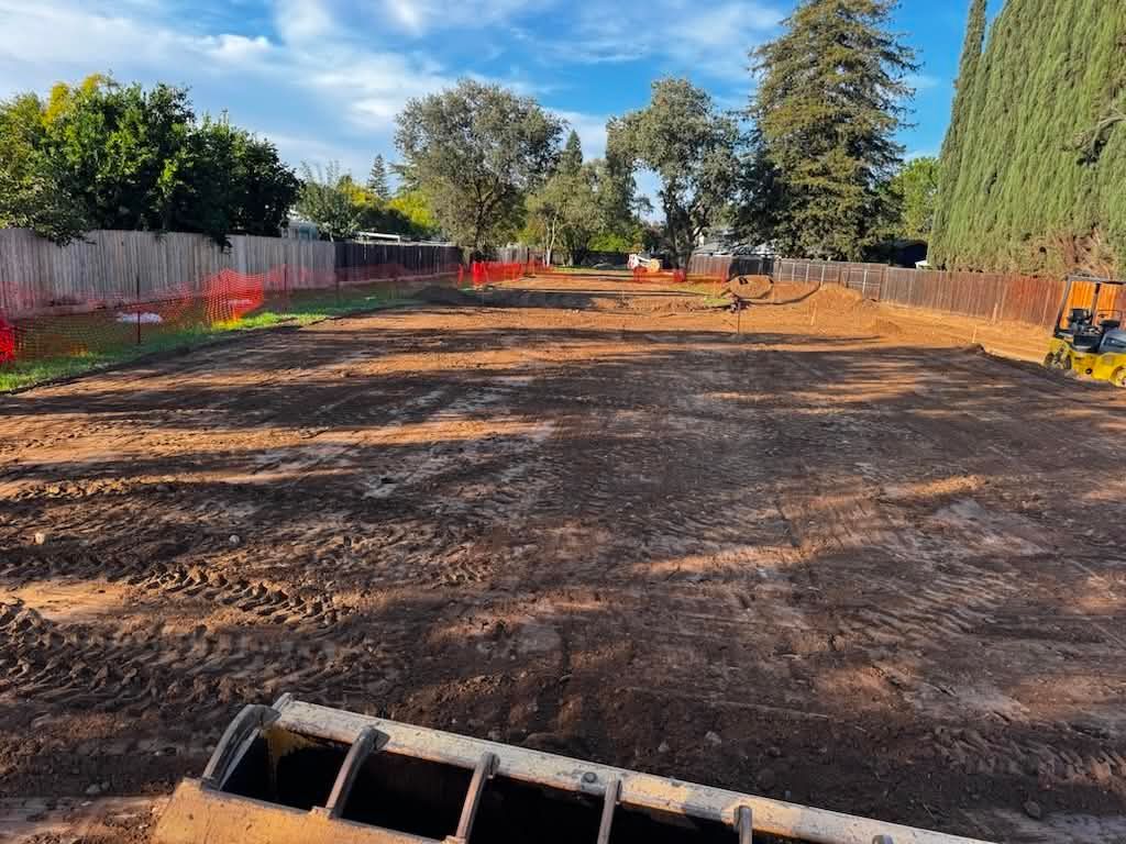 Cleared dirt lot with orange fencing and trees under a bright blue sky