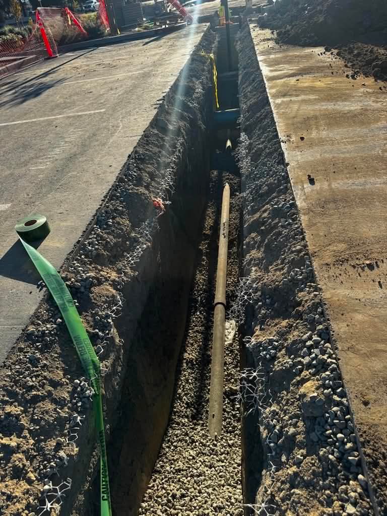 Open trench with exposed utility pipe beside a road construction area, marked by orange cones.