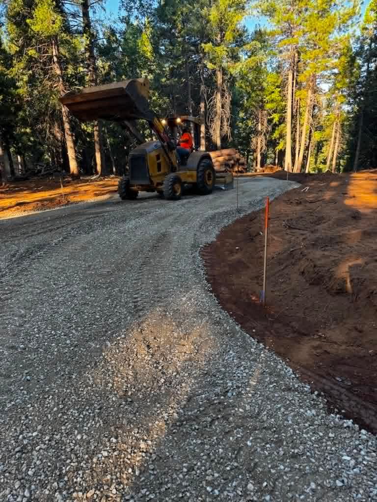 Backhoe dumping gravel on a winding dirt road in a wooded area