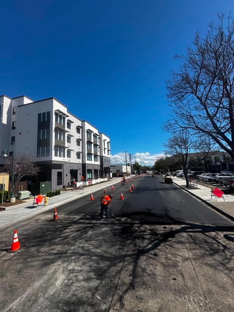 Road construction with orange cones beside a modern apartment building under a clear blue sky
