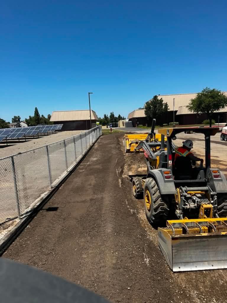 Construction equipment grading dirt along a fenced path beside a school building under a clear blue sky