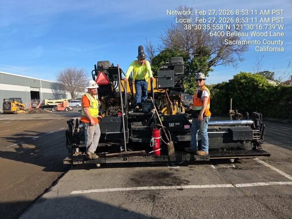 Road paving crew beside a black asphalt machine in a sunny parking lot, with traffic cones and safety vests.