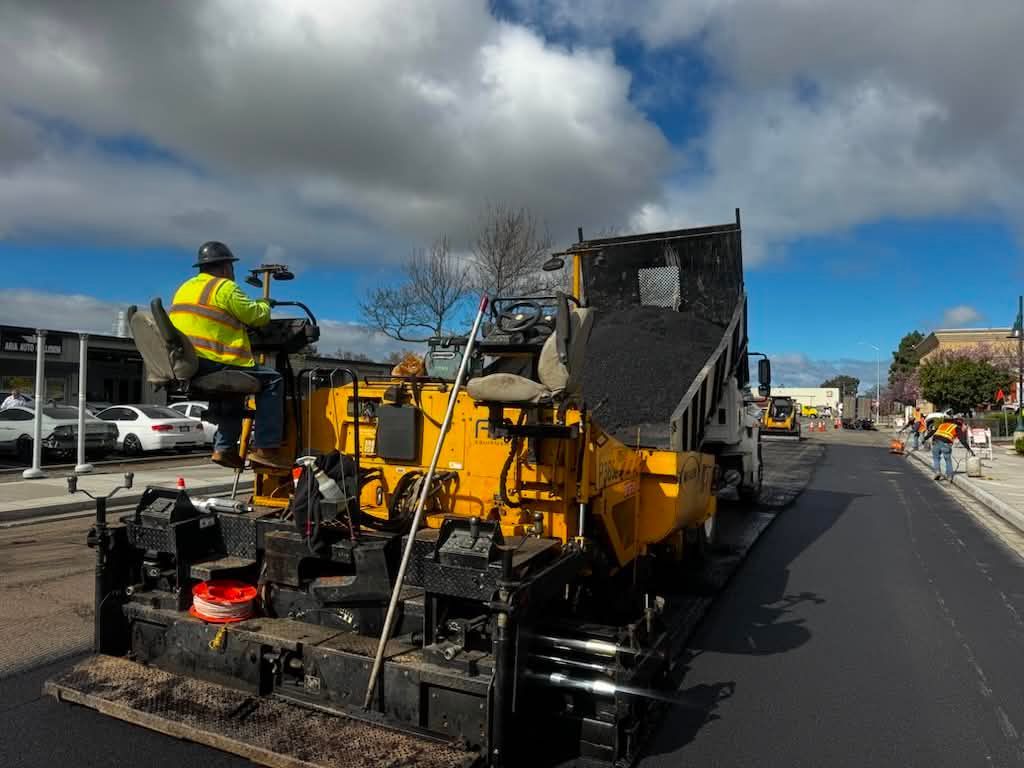 Road paving machine with workers in high-visibility gear on a street under cloudy sky