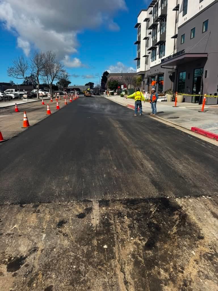 Road paving work with cones, workers, and a partially resurfaced street beside buildings under a blue sky
