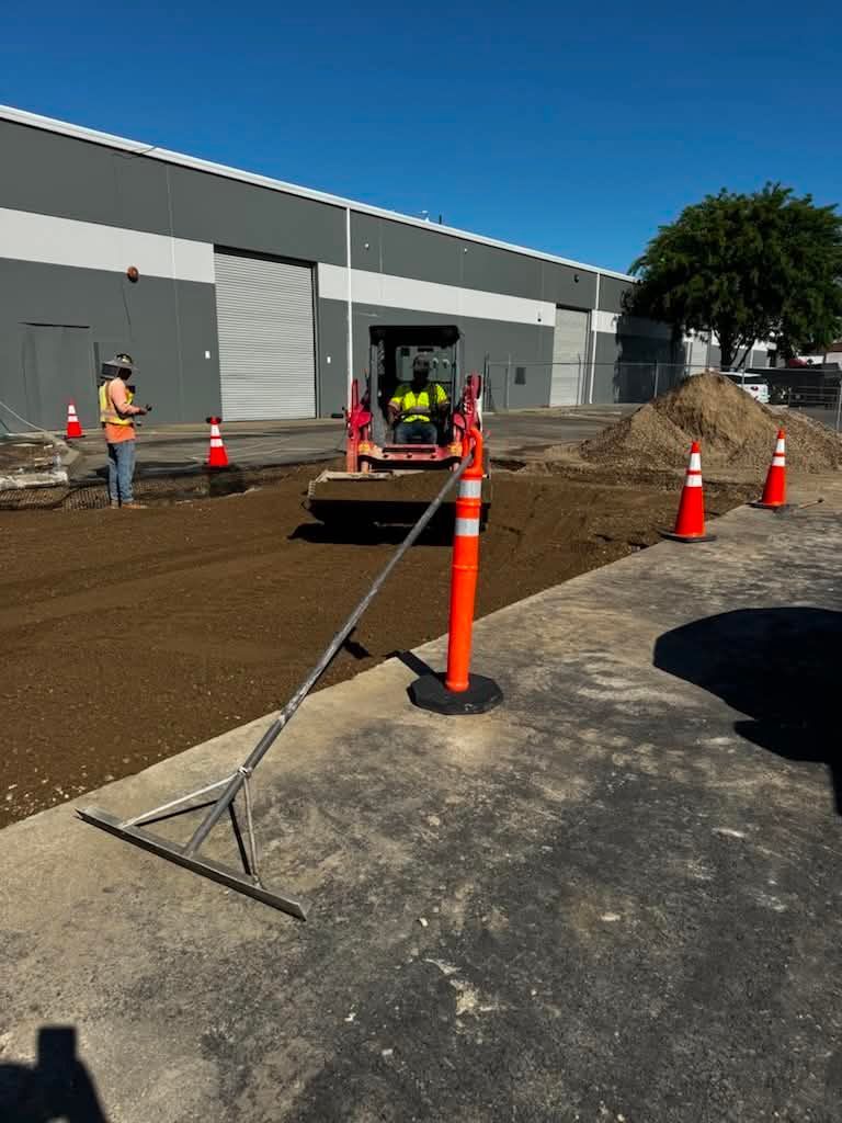 Construction workers and orange cones along a dirt trench beside a gray building under a blue sky