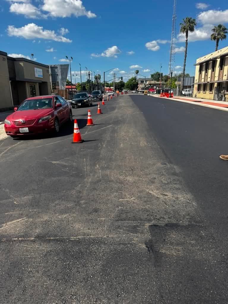 Roadwork on a sunny street with orange cones, parked red car, and buildings lining both sides
