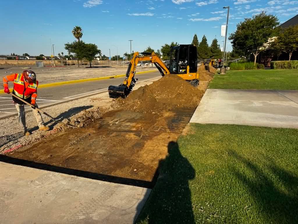 Roadwork with excavator digging a trench beside a sidewalk and grassy verge on a sunny day