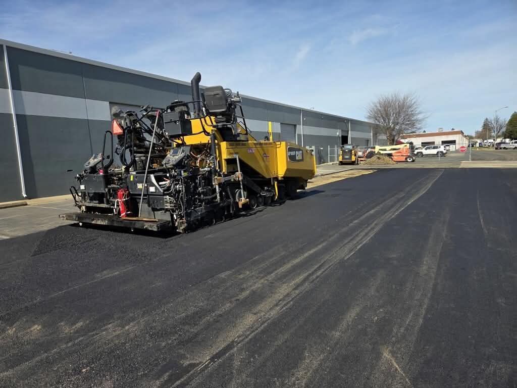 Paving machine lays fresh black asphalt beside an industrial building on a sunny day