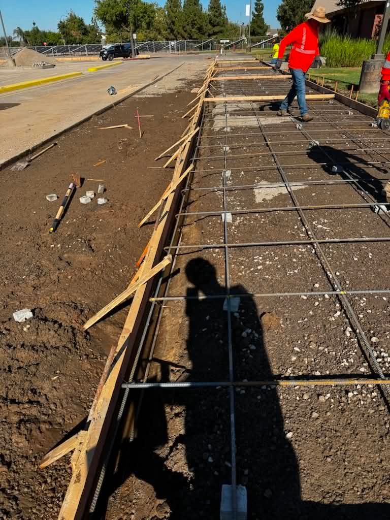 Roadside concrete work with rebar and wooden forms; worker in red vest stands on the right.