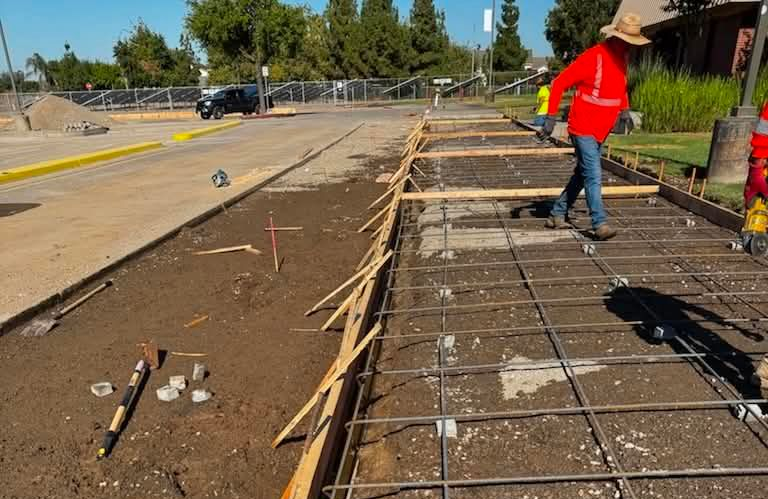 Roadside concrete work with rebar and wooden forms; worker in red vest stands on the right.