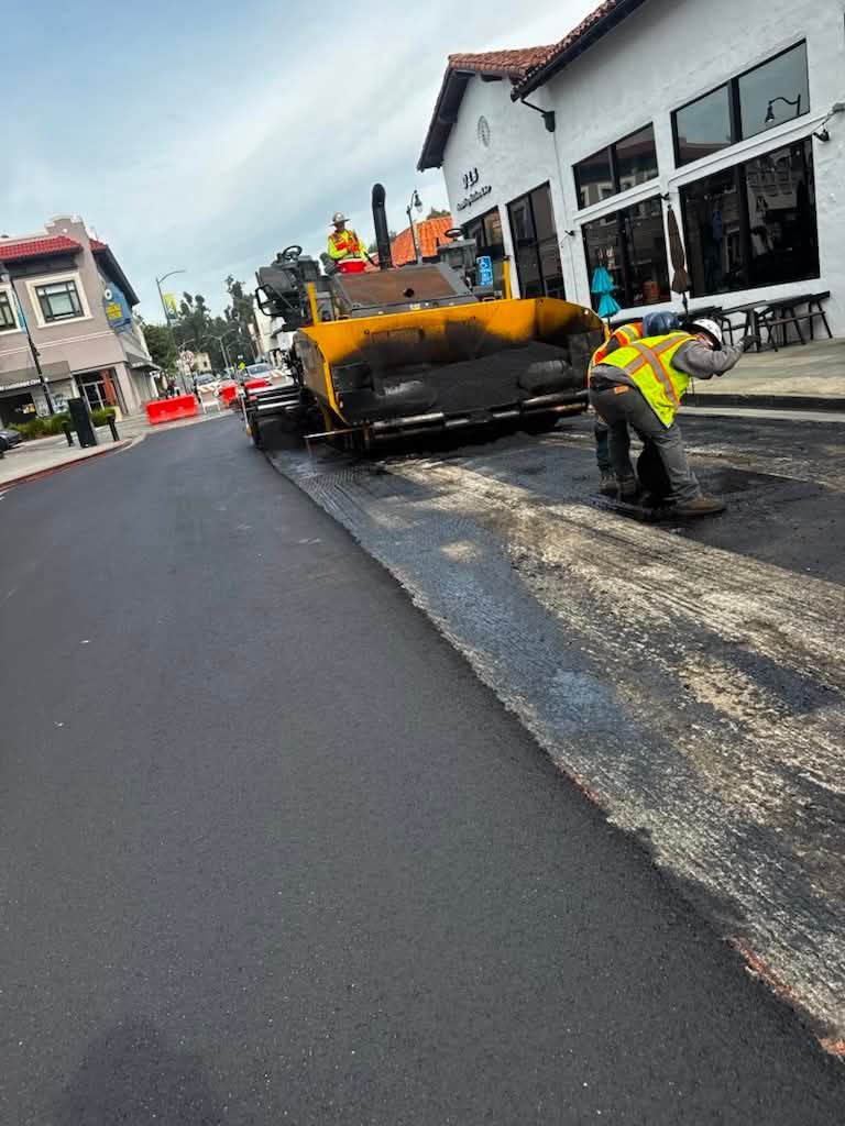 Road resurfacing with a yellow asphalt paver and worker on a wet street beside buildings