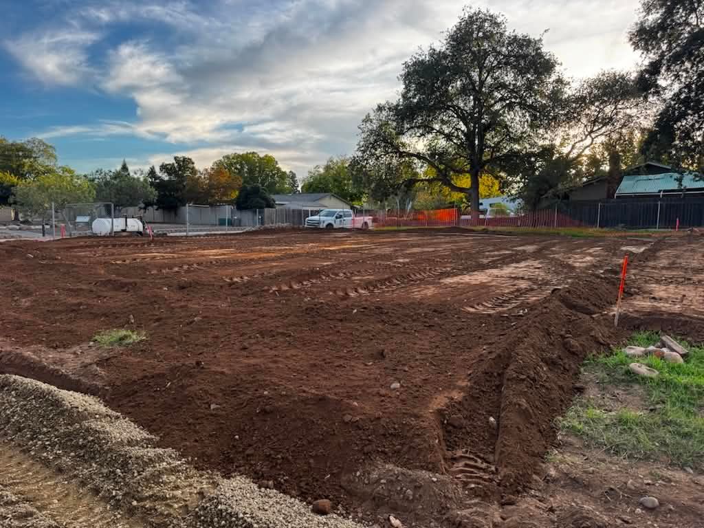 Cleared dirt lot with fresh grading, trees, and houses under a cloudy sky