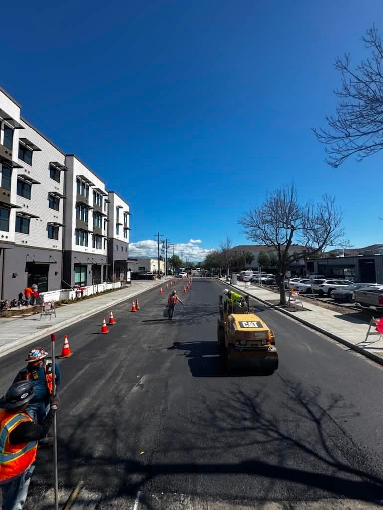 Road construction with cones, a worker, and a yellow utility truck on a city street beside apartments.